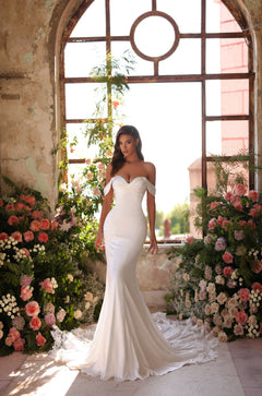 A bride in an off-shoulder white mermaid wedding gown poses among pink and white flowers by an arched window.
