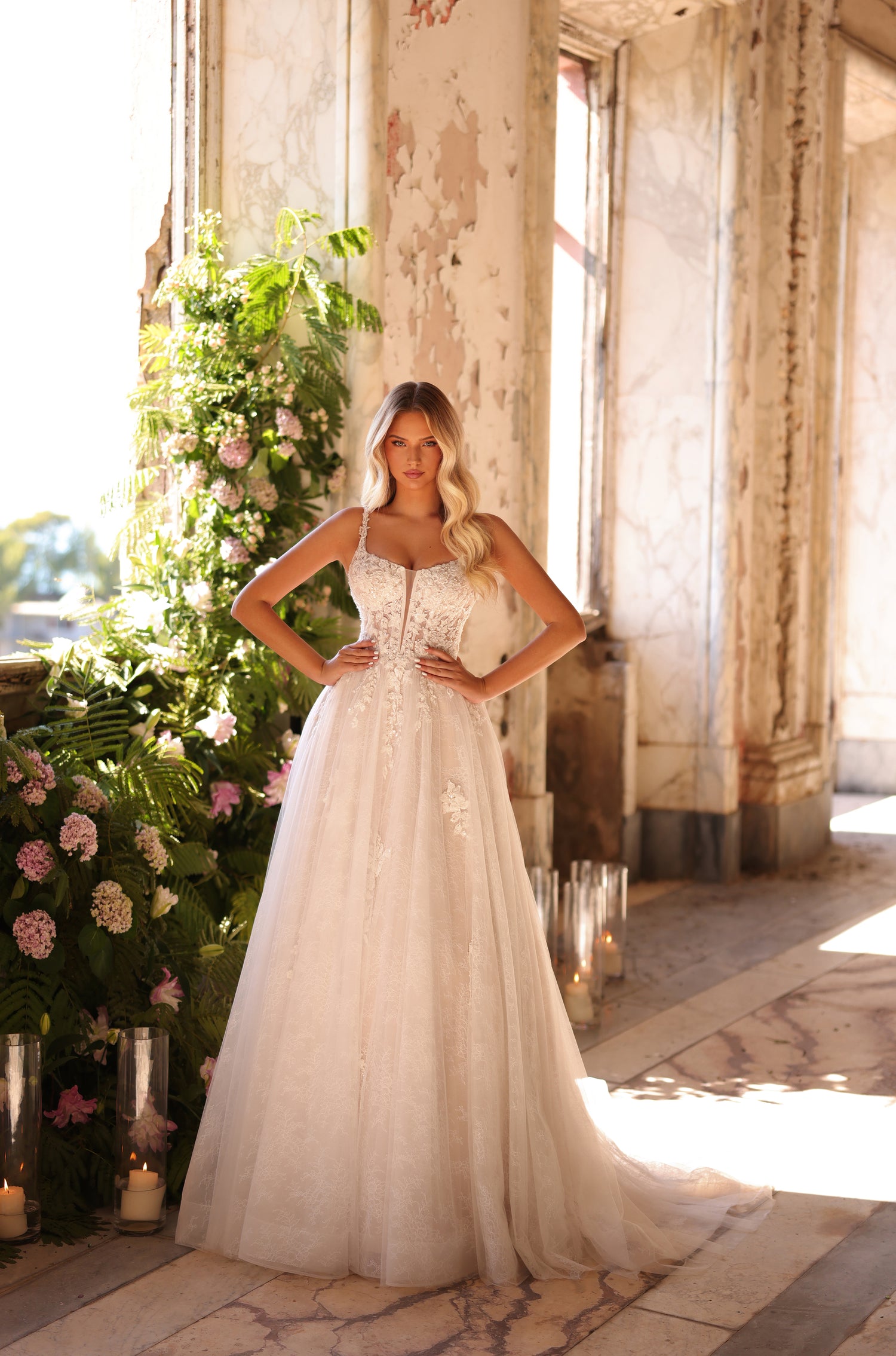 A bride in a white lace ballgown stands confidently in a sunlit, weathered room with floral decorations.