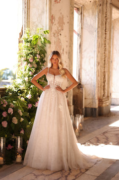 A bride in a white lace ballgown stands confidently in a sunlit, weathered room with floral decorations.