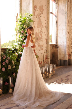 A bride in a lace A-line wedding dress stands by a window with flowers and candles, her long wavy hair catching the light.