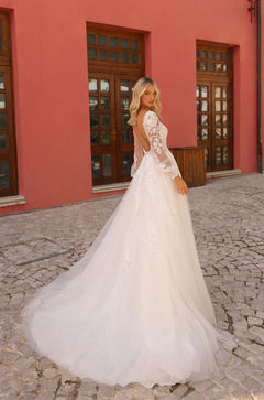 A bride in a lace long-sleeve white ballgown stands on cobblestone, with a salmon-colored building behind her.