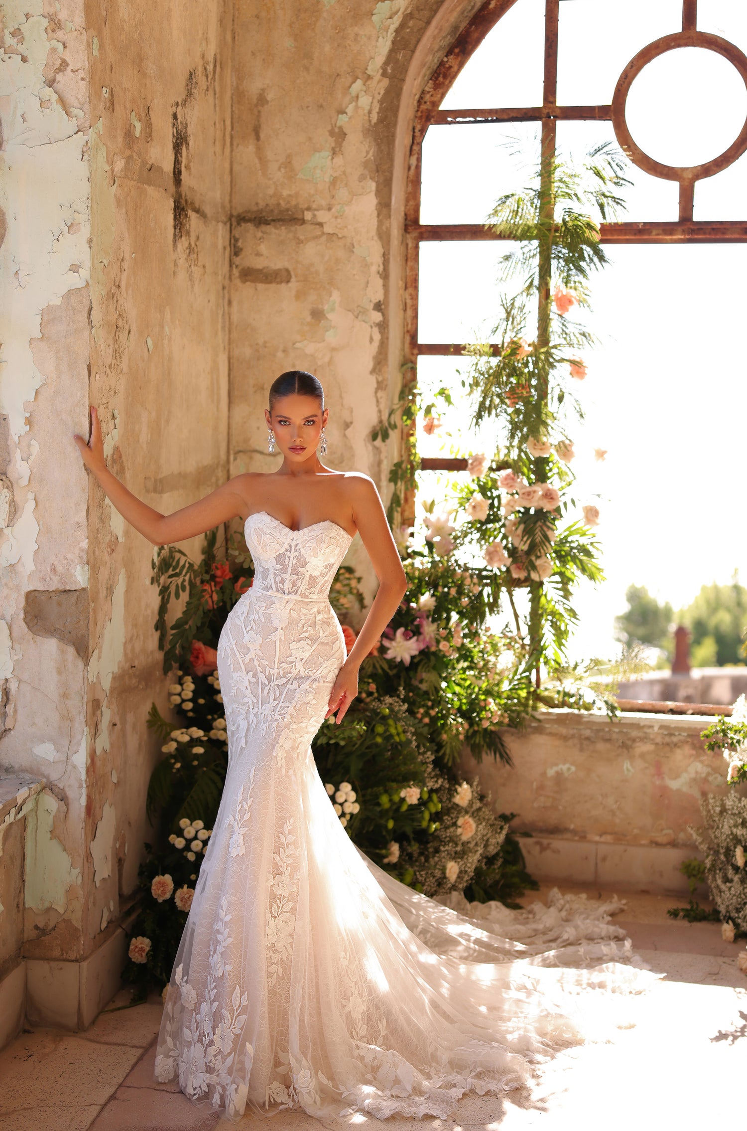 A bride in a strapless lace mermaid wedding gown poses in a sunlit, flower-filled rustic room.