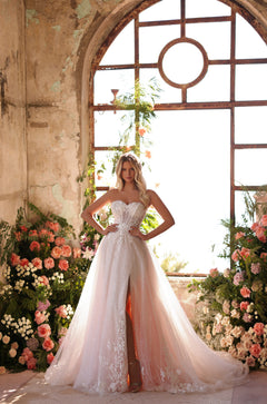 A bride in a sparkling strapless ballgown stands amid coral flowers by an arched, weathered window.