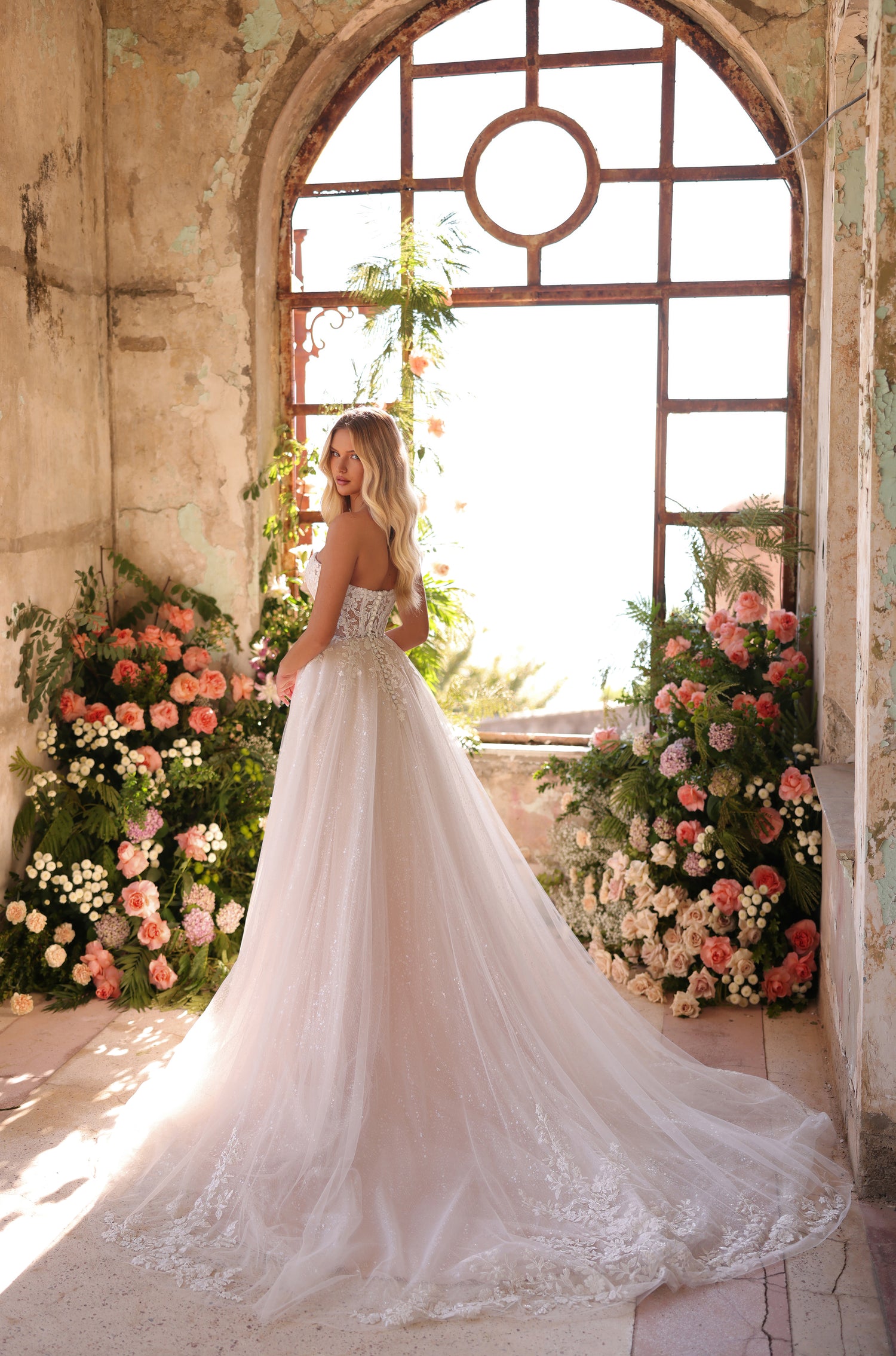 A bride in a lace ballgown stands by an arched window, surrounded by coral and pink floral arrangements.
