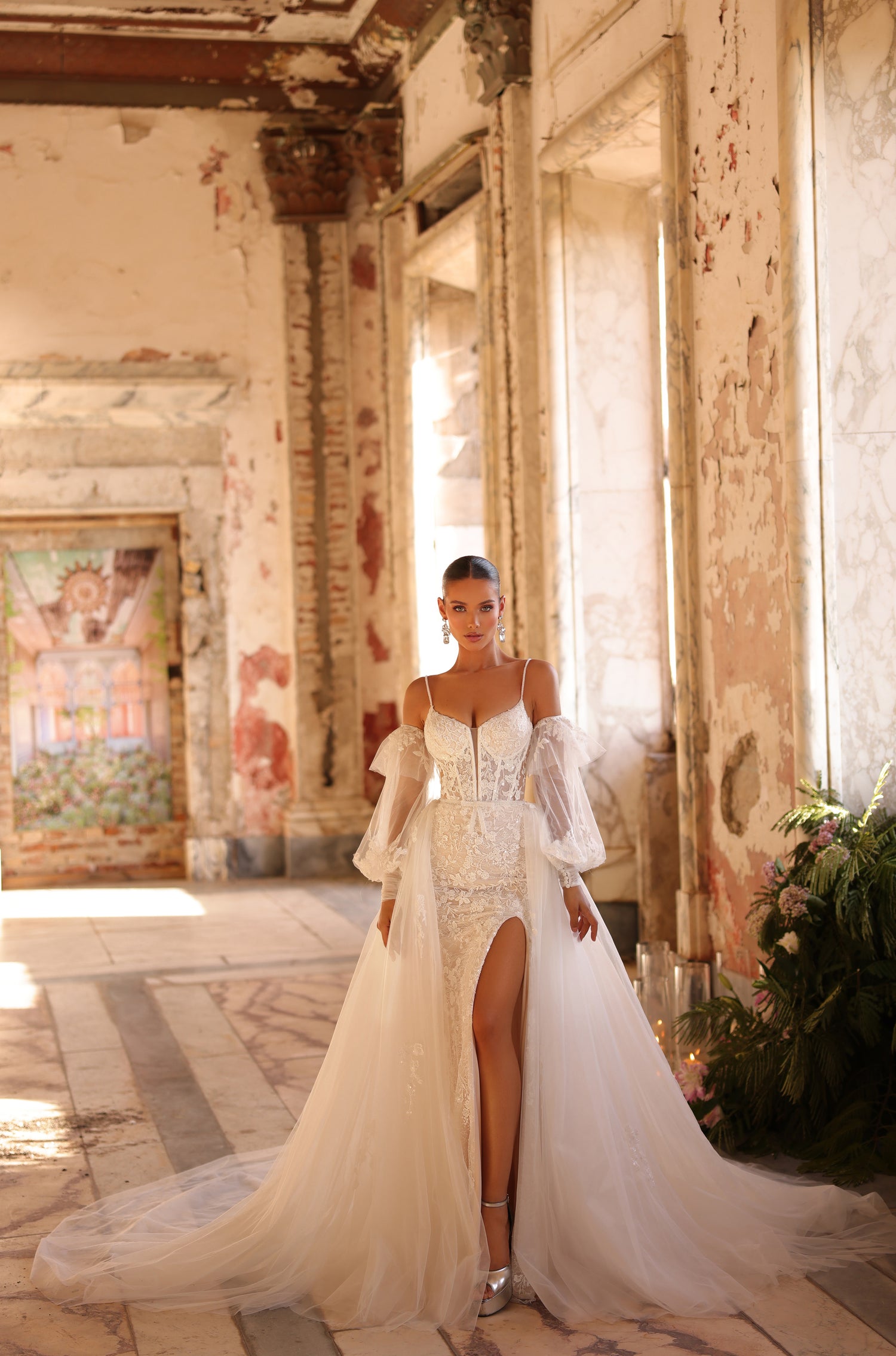A bride in an ornate white wedding gown with puffy sleeves stands in a distressed, sunlit historic room.