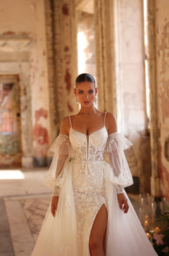A bride in an intricate lace wedding gown with off-shoulder sleeves stands in a sunlit, weathered interior.