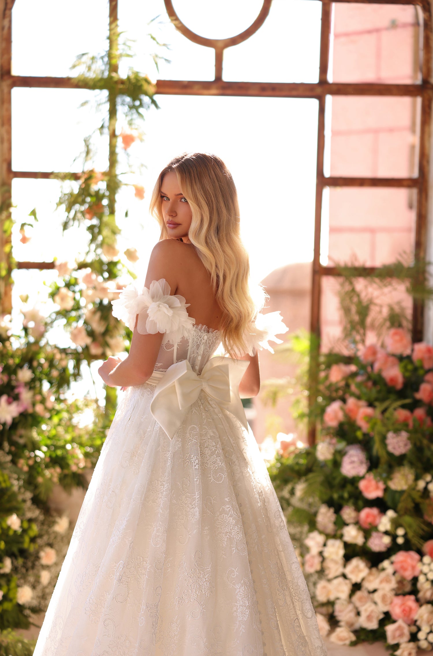 A bride in an off-shoulder lace wedding gown poses amid flowers by a vintage window.