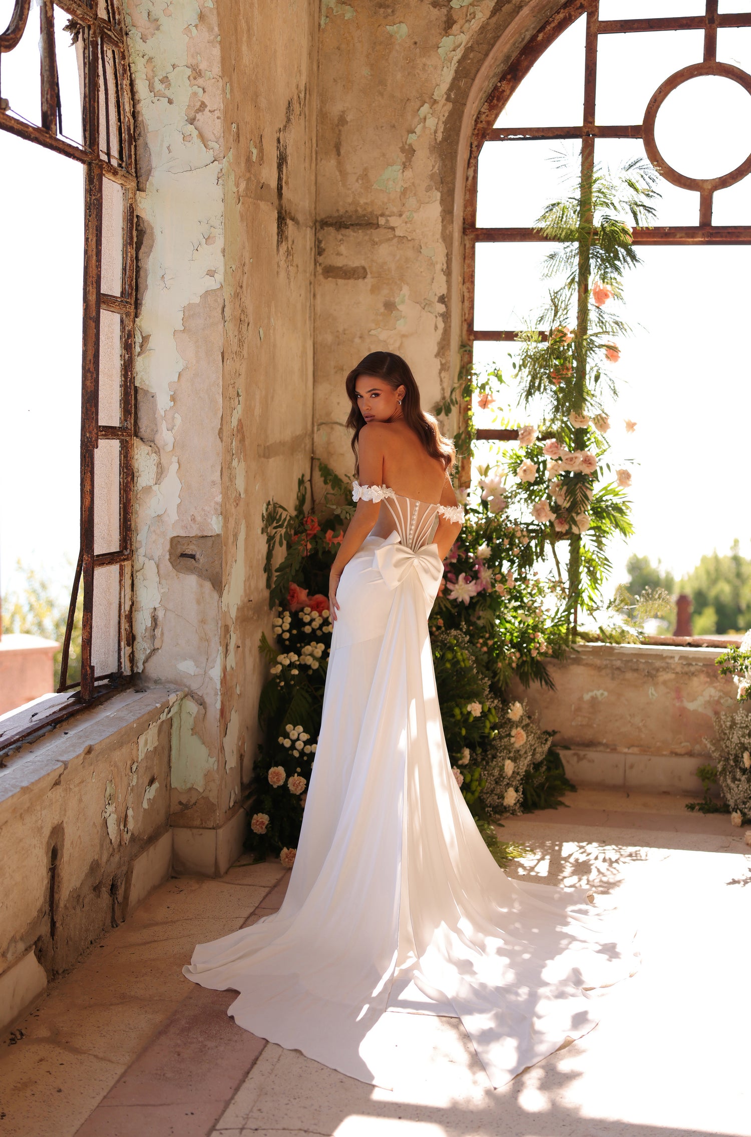 A bride in an off-shoulder white gown stands in a rustic, sun-drenched room with floral decorations and distressed walls.