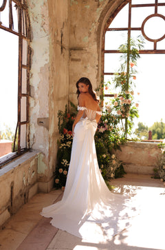 A bride in an off-shoulder white gown stands in a rustic, sun-drenched room with floral decorations and distressed walls.