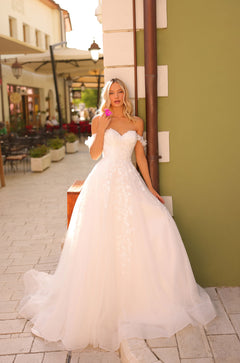 A bride in a strapless white lace ballgown stands on a cobblestone street with a pink flower.