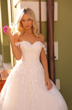 A blonde woman in an off-shoulder white lace wedding gown holds a pink flower against an olive green backdrop.