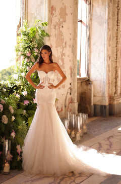 A bride in a strapless mermaid wedding gown poses in a sunlit, distressed room with floral and candle decor.