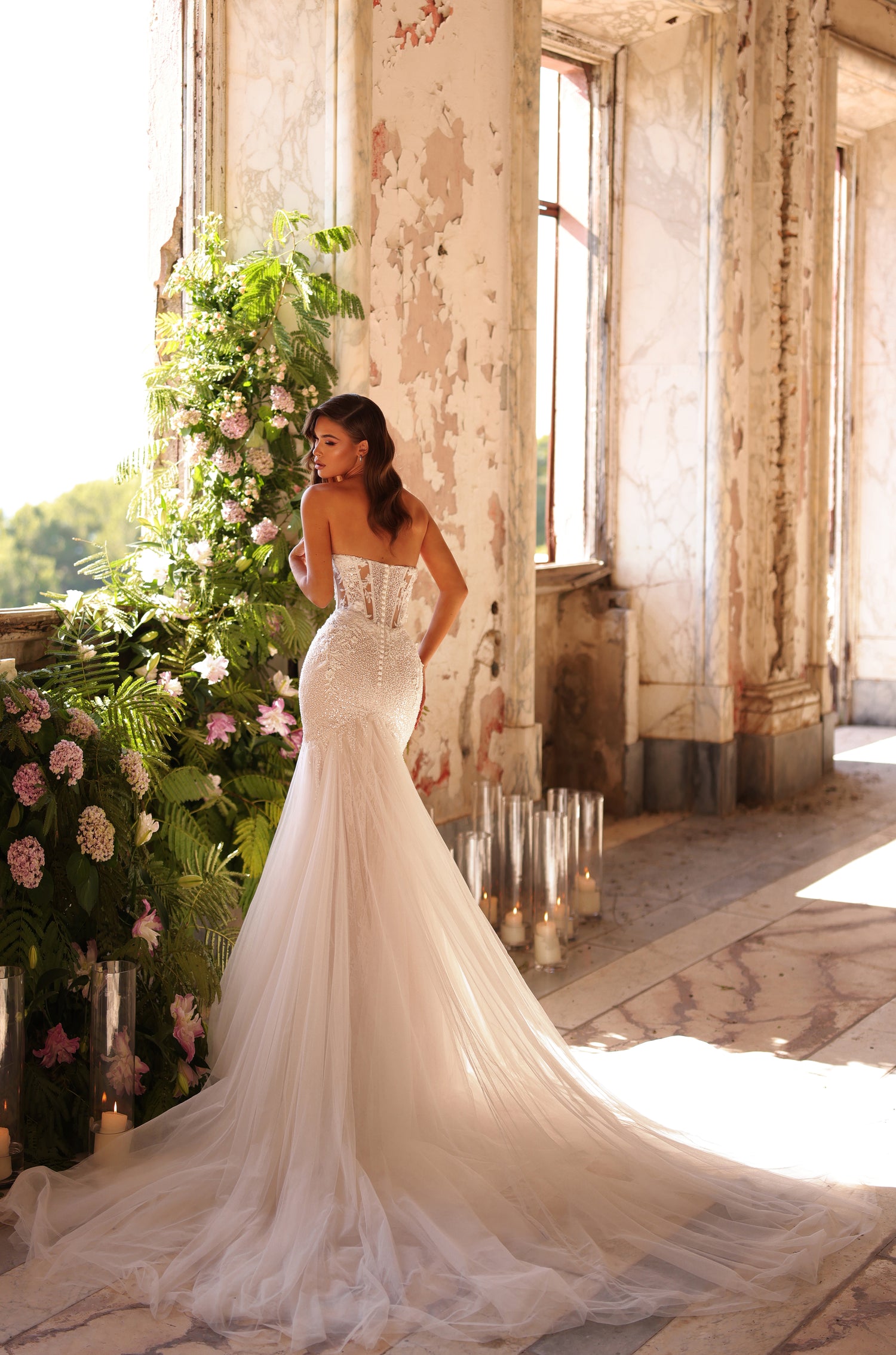 A bride in a strapless mermaid wedding gown stands in a sunlit, weathered room with floral and candle decor.