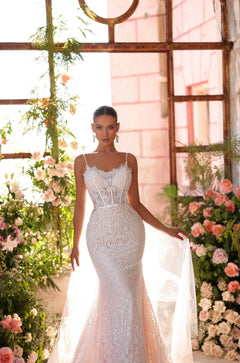 A bride in an intricate white lace mermaid wedding gown stands amid blush roses in a sunlit greenhouse.
