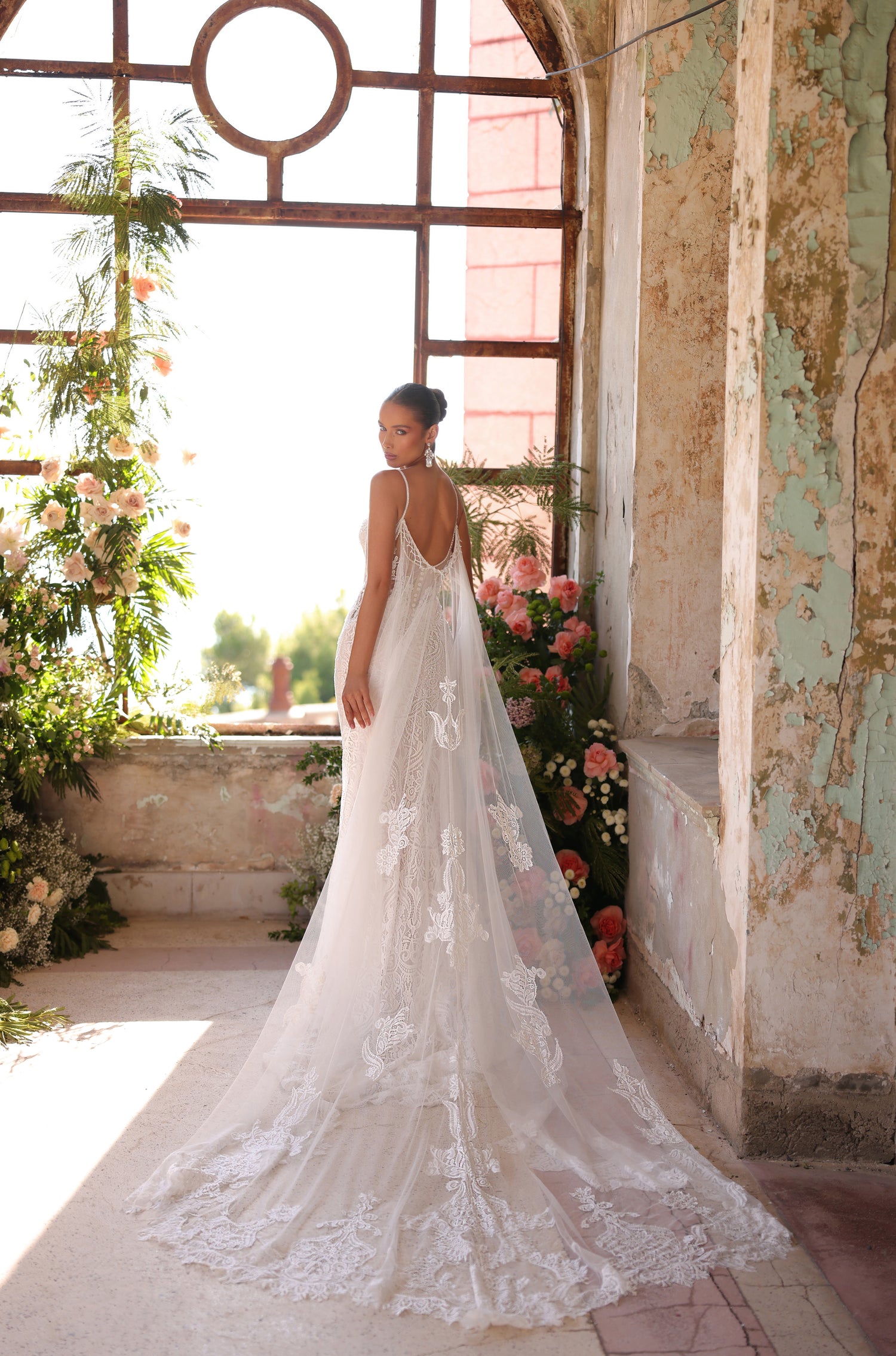 A bride in a delicate lace wedding gown stands by a window with floral arrangements in a weathered, vintage room.
