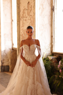 A bride in a detailed lace off-shoulder wedding gown with flowing veil stands in a soft-lit, vintage room.
