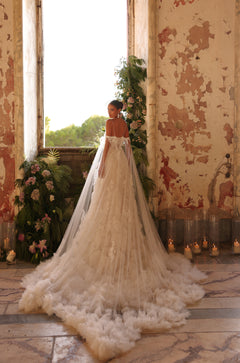 A bride in an ornate lace gown with a long train stands by a window with peeling walls and floral decor.
