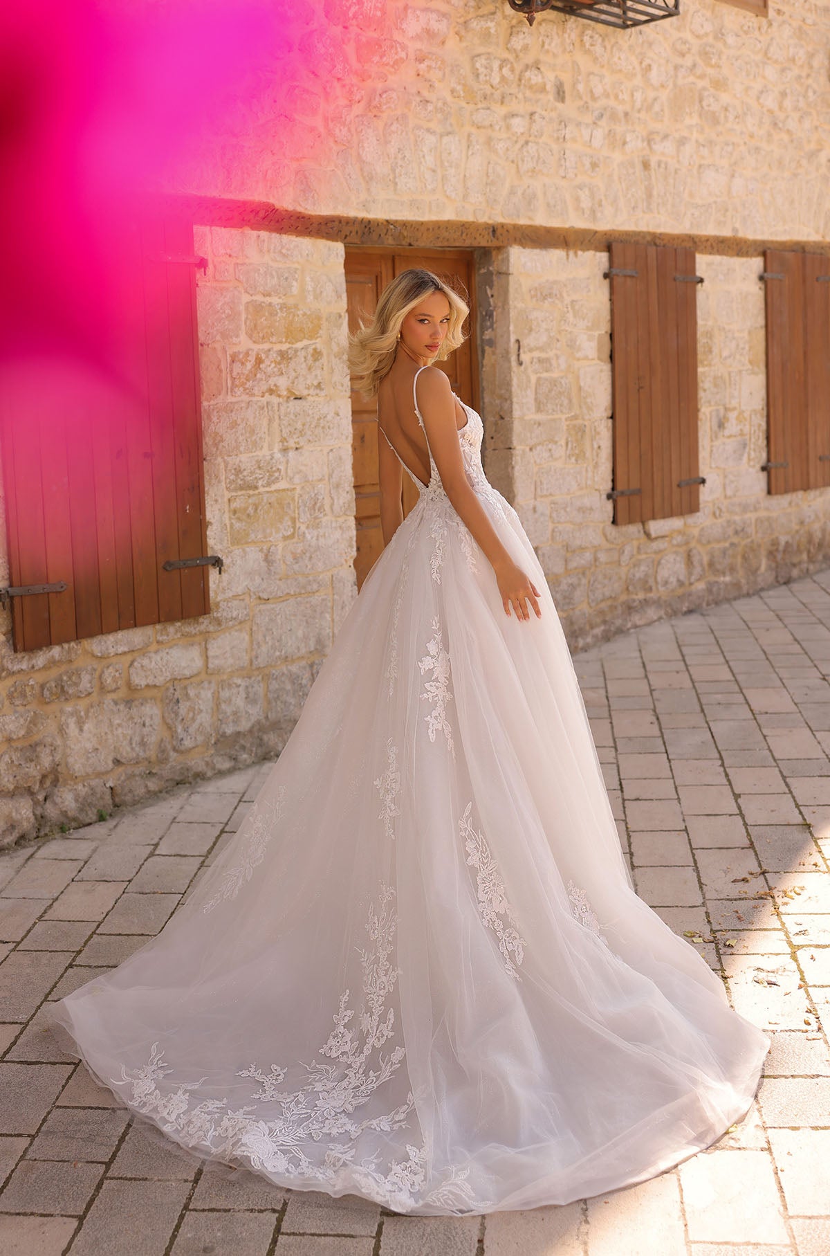 A bride in a white lace ballgown with an open back poses by a stone wall with wooden shutters.