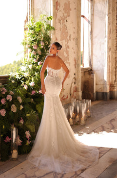 A bride in a strapless lace mermaid wedding gown stands in a sunlit, distressed room with floral decor and candles.