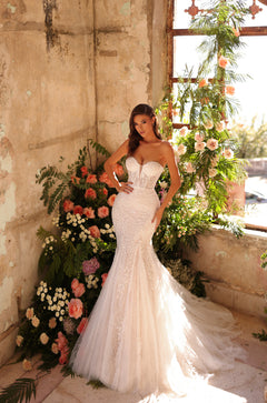 A bride in a white mermaid-style lace wedding dress poses amid coral roses and green foliage in a rustic setting.