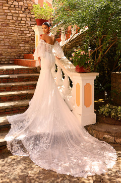 A bride in an off-shoulder lace wedding gown stands on stone steps with a flowing train amid greenery.