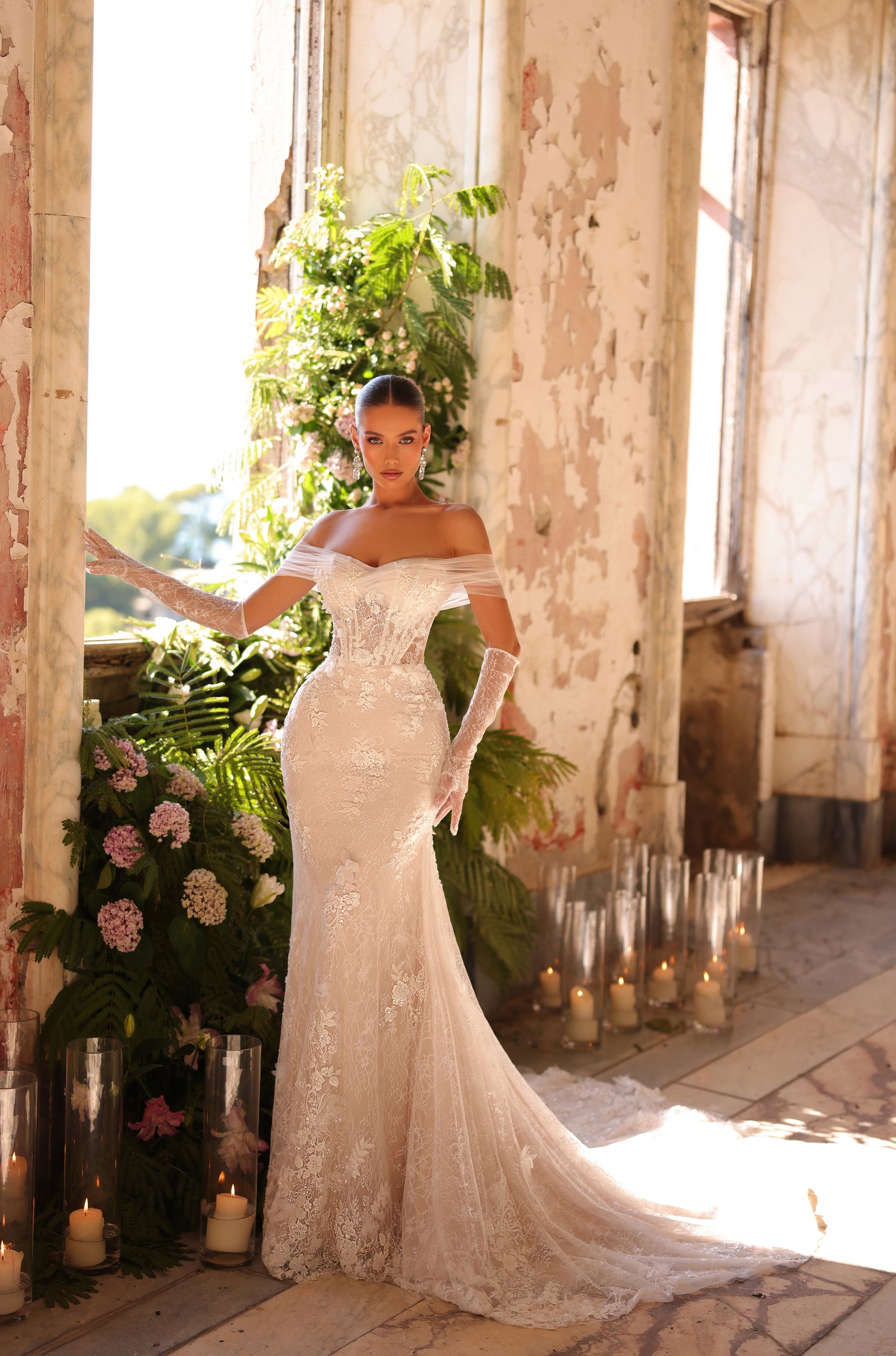A bride in an off-shoulder lace mermaid wedding gown poses amid candles and lush greenery in a vintage room.