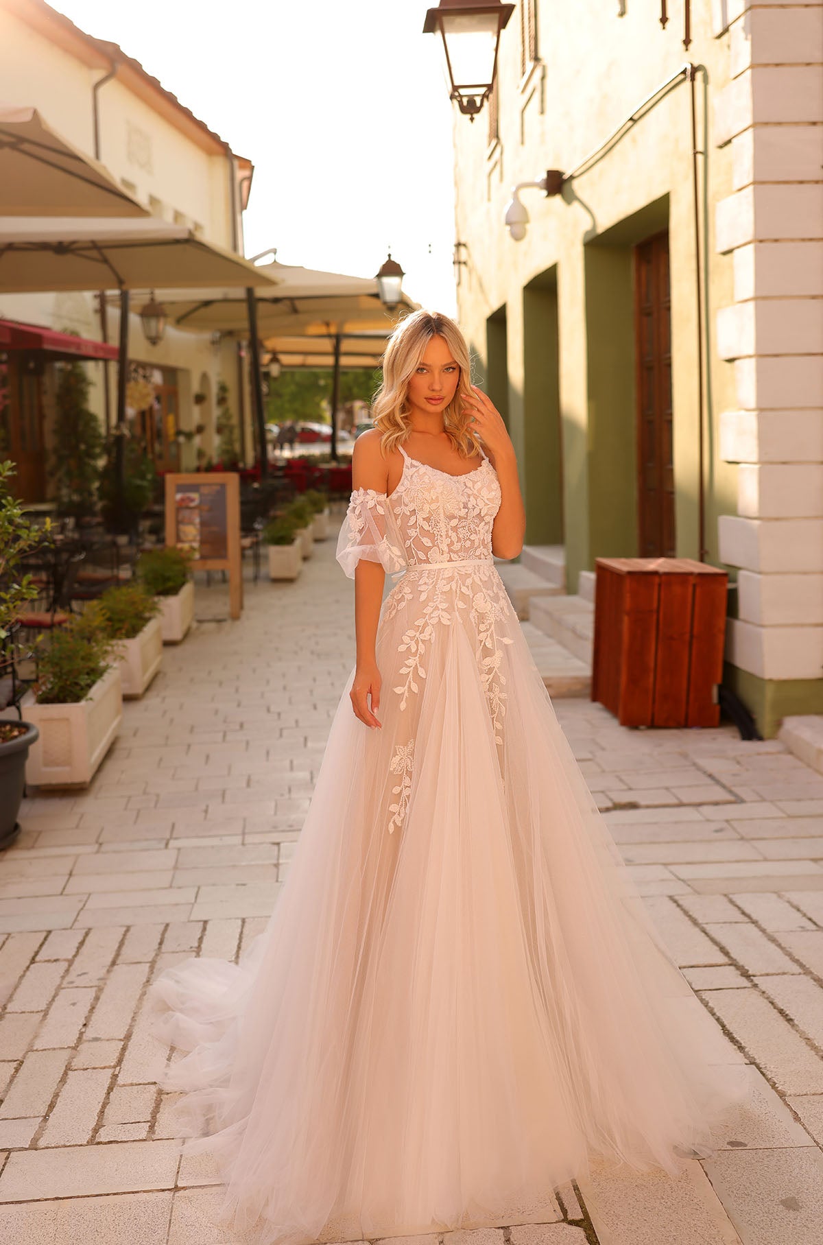 Blonde woman wears off-shoulder lace wedding gown with flowing tulle skirt in European street setting.