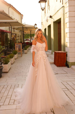 Blonde woman wears off-shoulder lace wedding gown with flowing tulle skirt in European street setting.