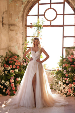 A bride in a flowing white lace corset gown stands amid pink roses by an arched rustic window.