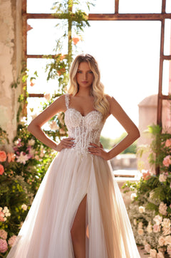 A bride wears a white lace corset wedding gown with a tulle skirt, posing in a floral-filled greenhouse.