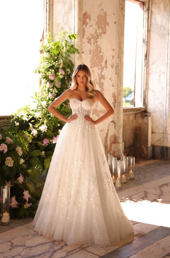 A bride in a strapless lace ballgown stands confidently in a sunlit, plant-filled room with peeling walls.