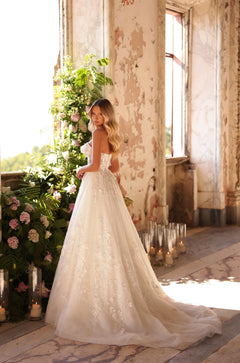 A bride in an elegant, lace-detailed white wedding gown stands in a sunlit, vintage room with floral and candle decor.