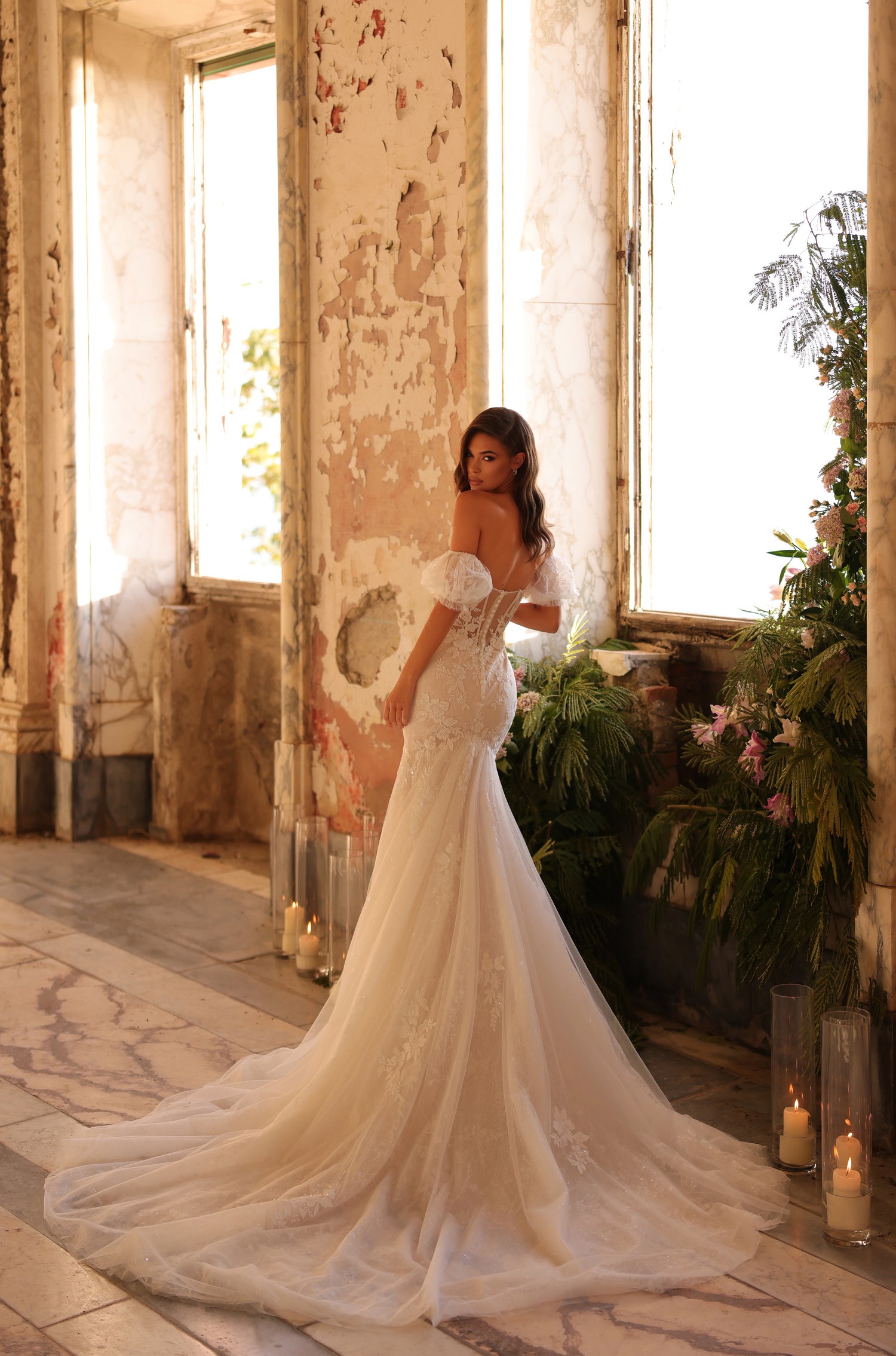 A bride in a lace mermaid wedding gown stands by a window with peeling walls and greenery.