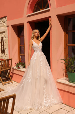 A blonde bride in a lace ball gown stands by a salmon-colored wall with arched windows and potted flowers.