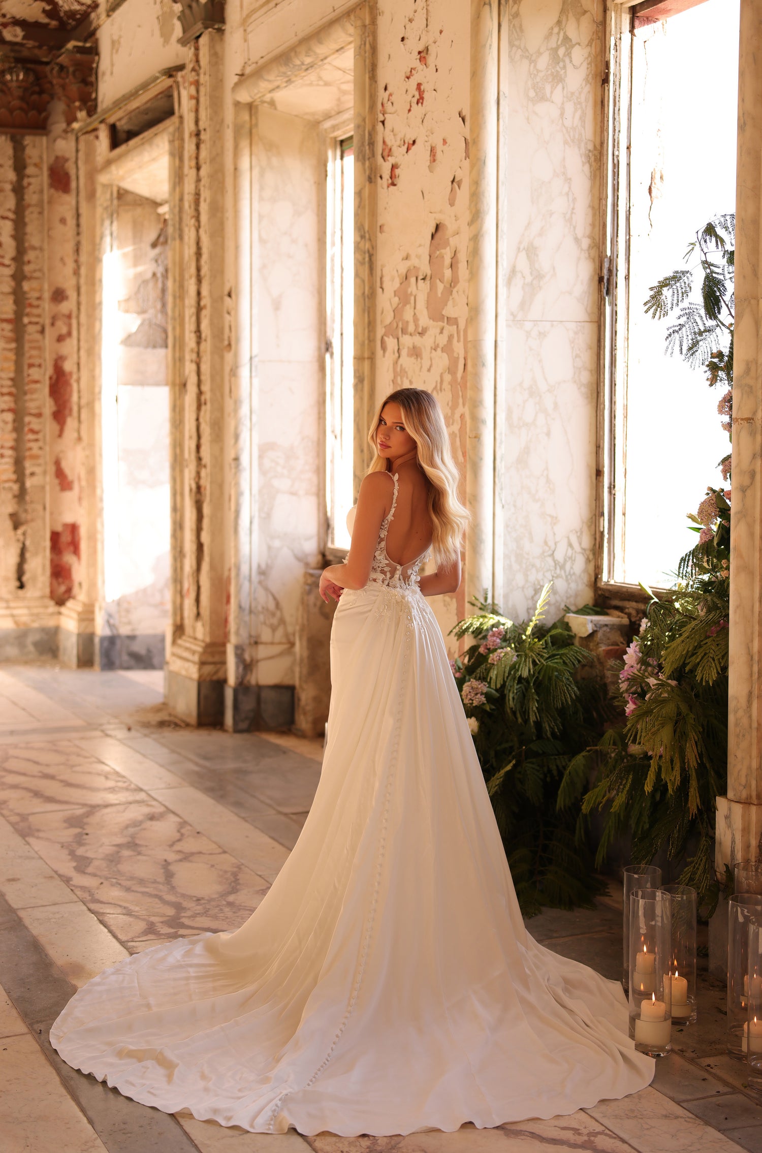 A bride in a flowing white gown stands by a window in a weathered, sunlit room with lush greenery.