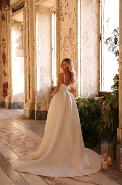 A bride in a white ball gown stands in a vintage room with plants and candles, facing away from the camera.