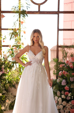 A blonde woman wears an elegant white lace wedding gown with a full skirt, standing among soft-colored flowers.