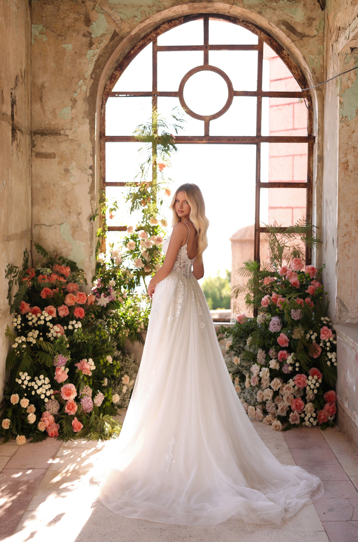 A bride in a white lace gown stands amid floral arrangements by an arched window in a rustic room.