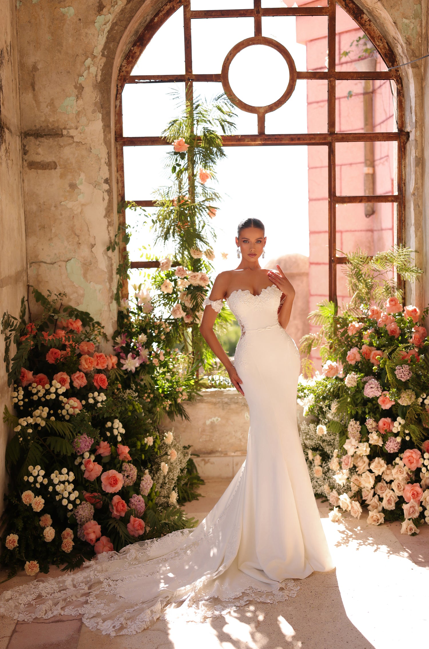 A bride in a white mermaid wedding gown stands amid lush pink and white floral arrangements by an arched window.