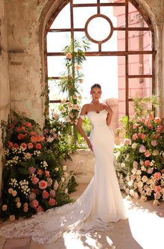 A bride in a white mermaid wedding gown stands amid lush pink and white floral arrangements by an arched window.