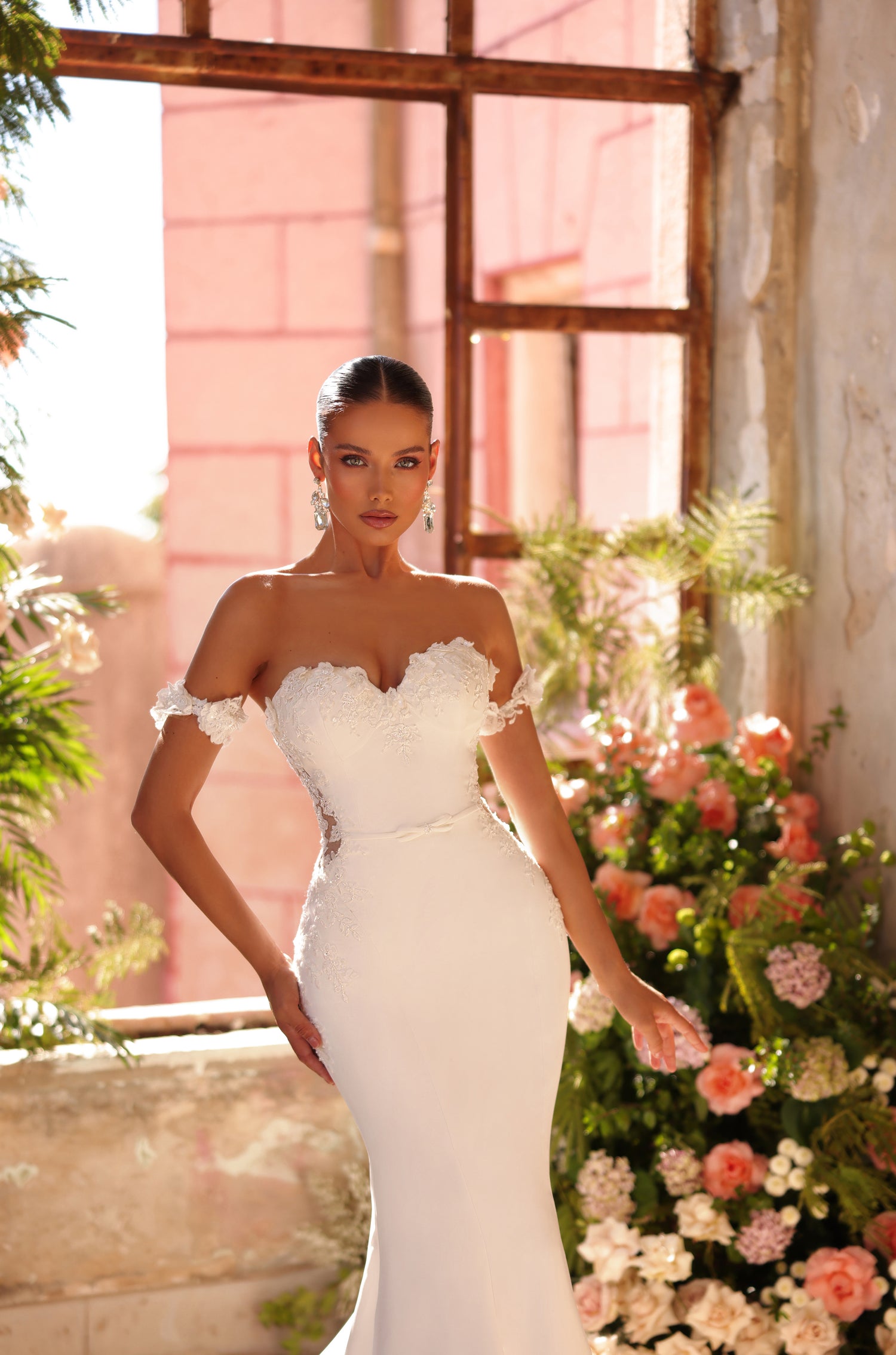A woman in an elegant white mermaid-style wedding dress poses amid soft pink flowers and rustic windows.