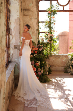 A bride in an off-shoulder lace wedding gown stands by a window with floral decor and peeling walls.