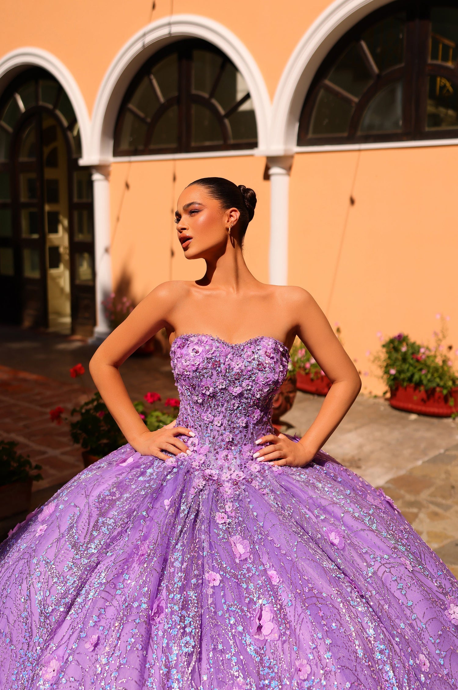 A person wears a lavender ballgown with intricate floral embellishments, posing in an arched courtyard.