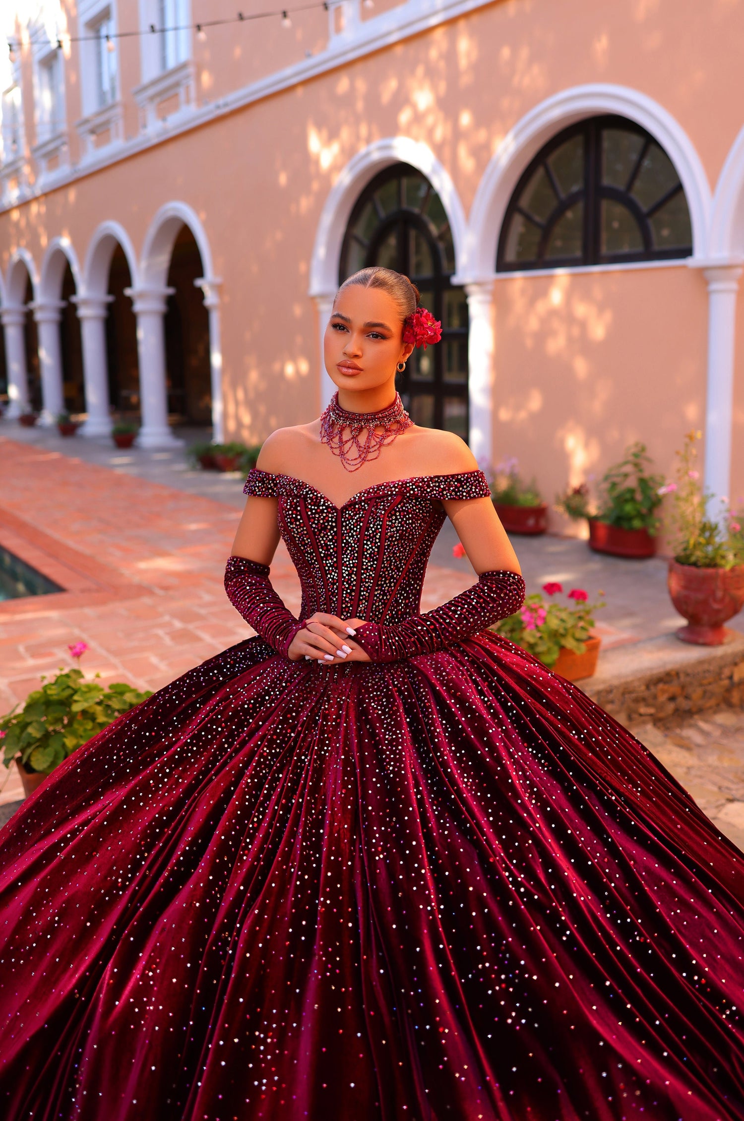 A woman wears a deep red sparkly ball gown with off-shoulder sleeves in an arched courtyard setting.