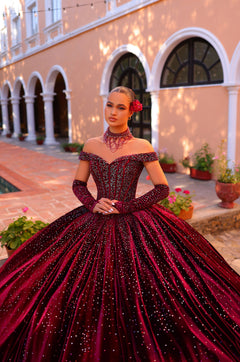 A woman wears a deep red sparkly ball gown with off-shoulder sleeves in an arched courtyard setting.