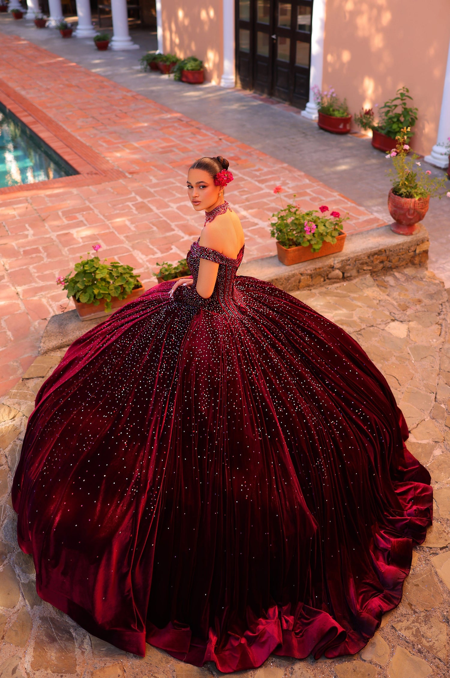 A teen wears a deep red sparkly ballgown with a massive skirt, posing near a pool and potted flowers.