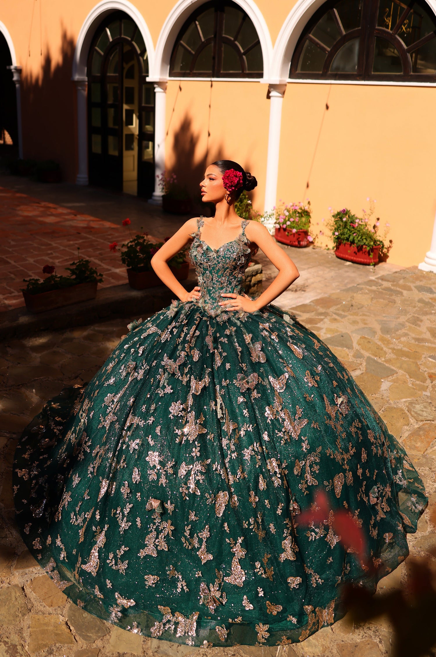 A woman wears a voluminous emerald/gold ball gown with floral pattern, standing in an arched courtyard.