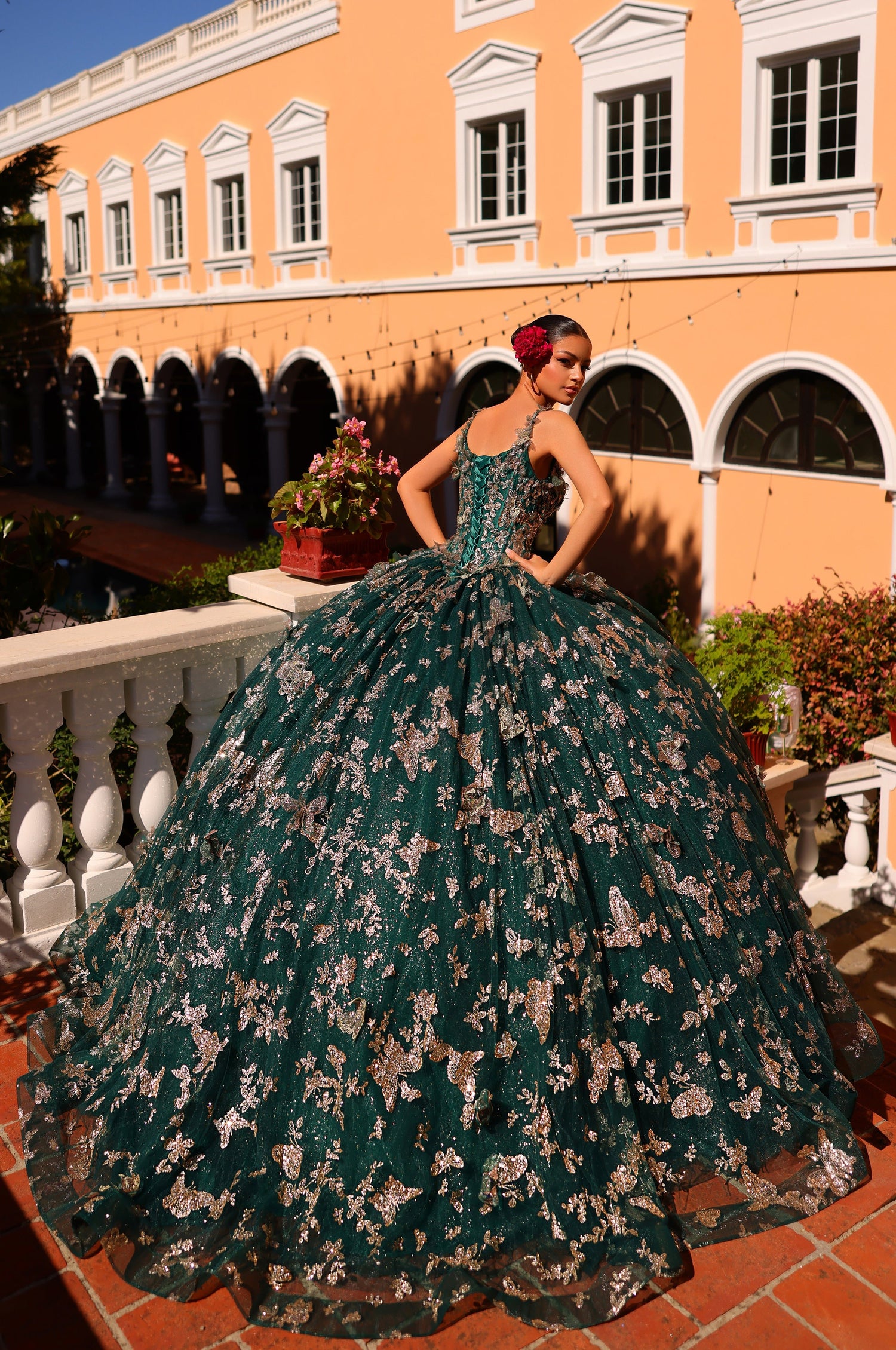 A woman in a deep green and silver butterfly-patterned ball gown poses in an ornate courtyard with arched windows.