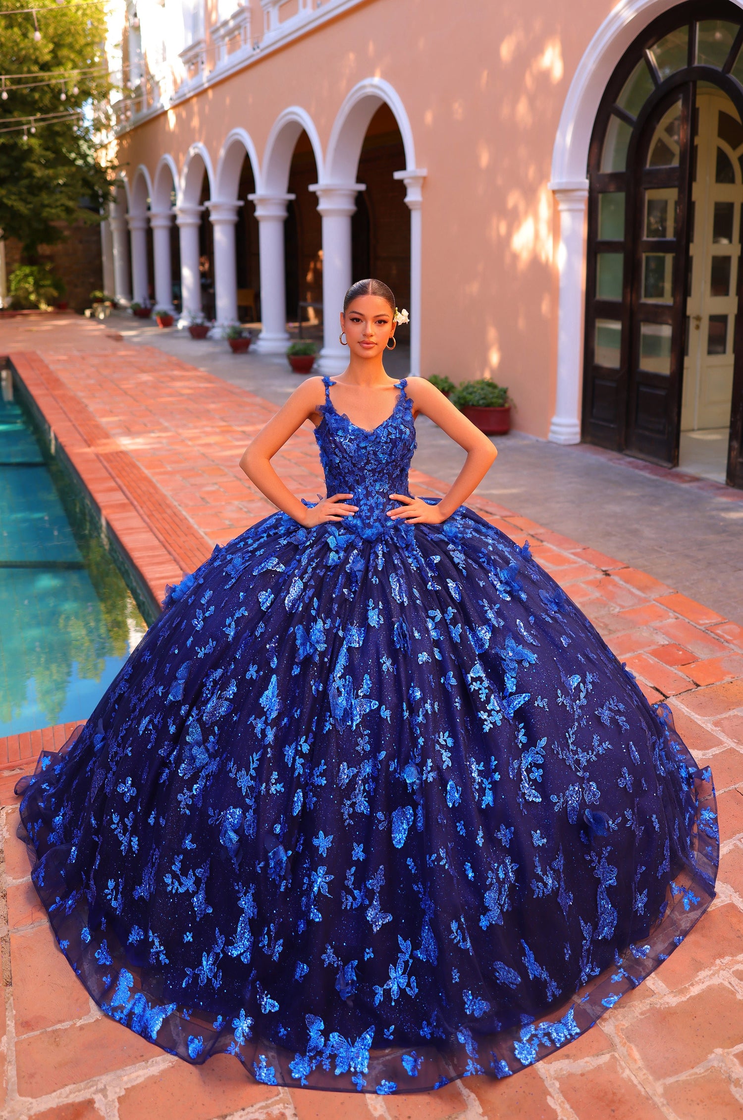 A young woman wears a voluminous Royal Blue ballgown with floral appliqués, standing by a tiled pool and columned building.