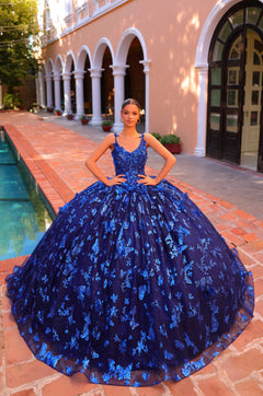A young woman wears a voluminous Royal Blue ballgown with floral appliqués, standing by a tiled pool and columned building.